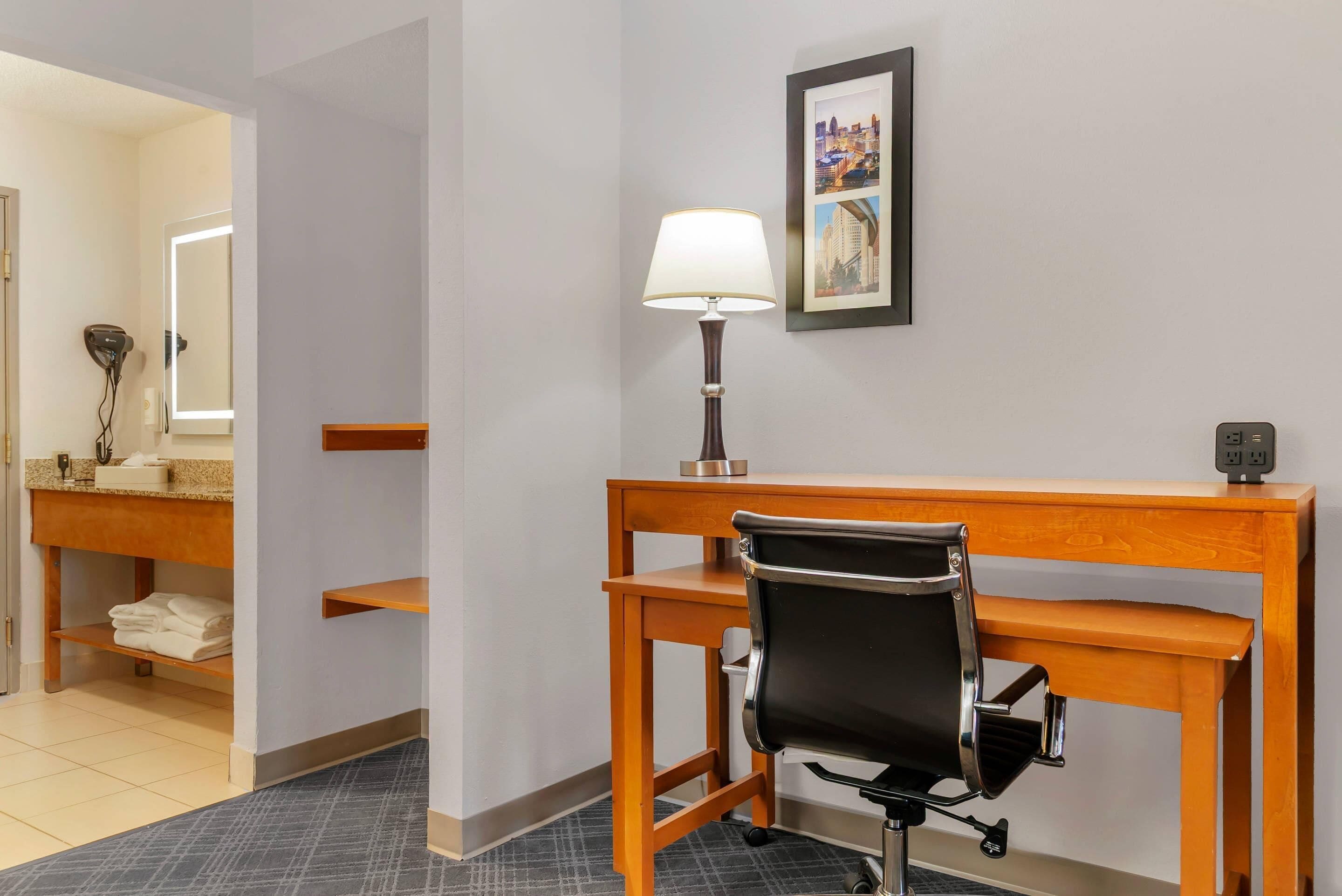 A hotel room with a wooden desk, a black office chair, a desk lamp, a framed cityscape picture, and a bathroom with a vanity and towel rack.