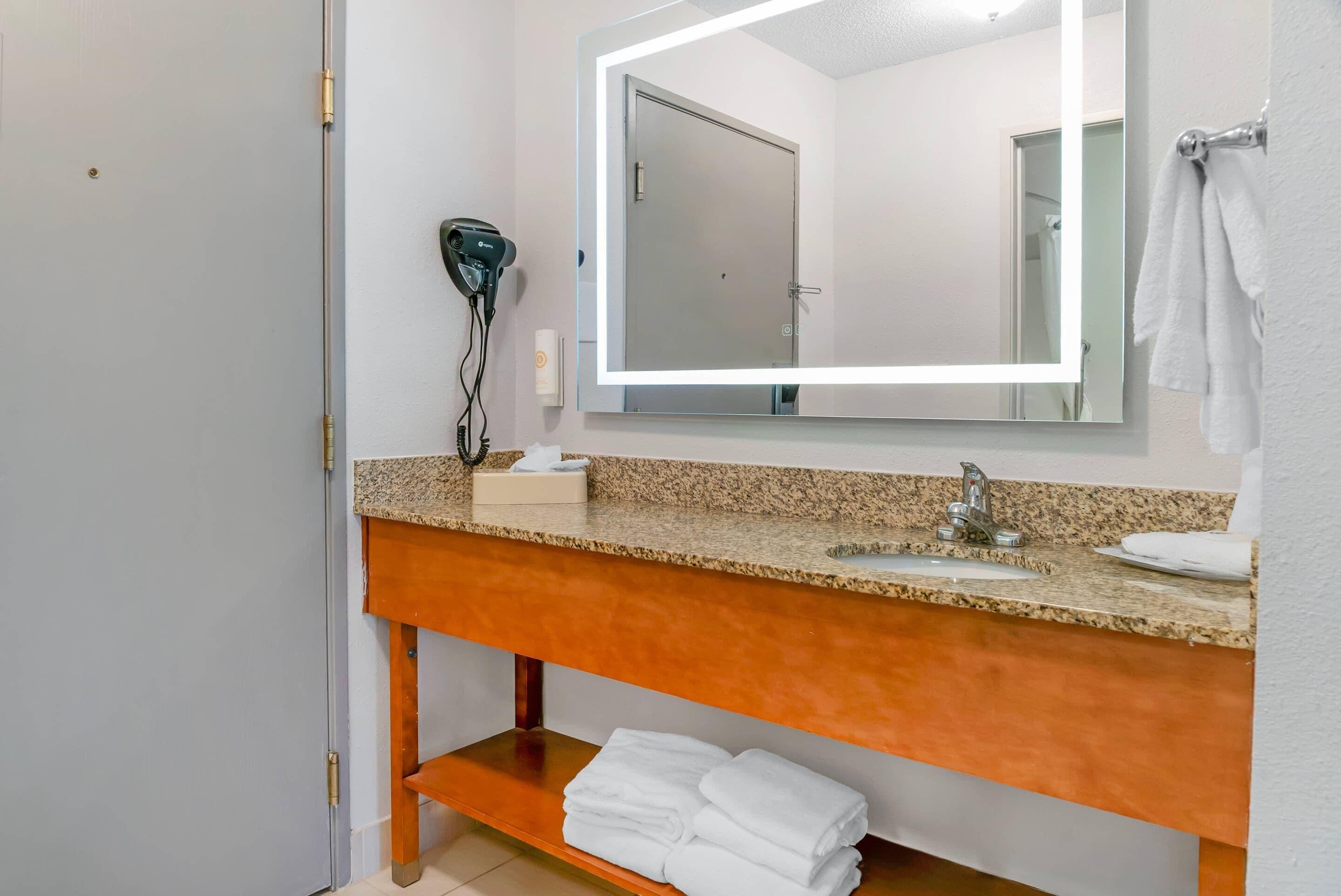 Un baño con lavamanos de encimera de granito, un espejo con iluminación integrada, un secador de pelo y toallas dobladas ordenadamente en un estante.