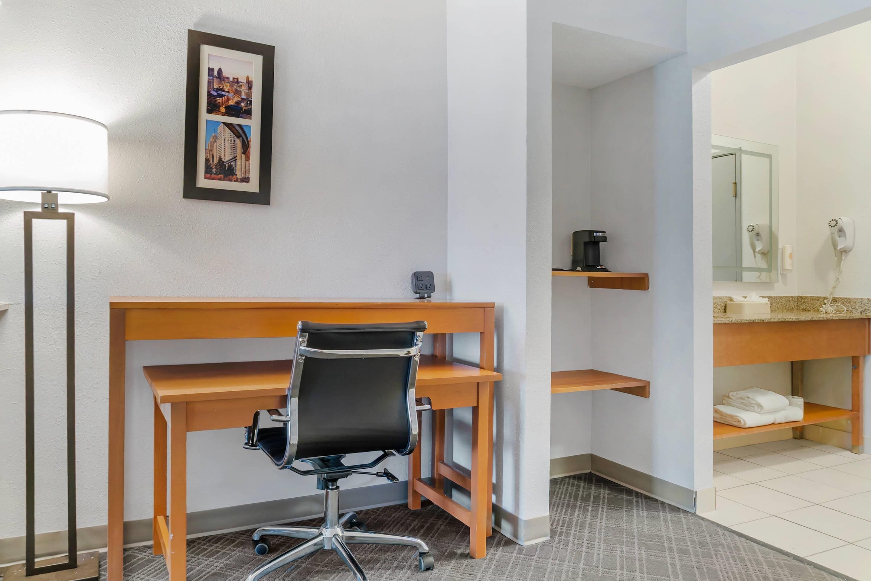 A hotel room with a wooden desk, a black office chair, a framed cityscape picture, a standing lamp, a coffee machine, a mirror, and a towel rack.