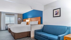 Habitación de hotel con dos camas, un sofá azul, una mesita de noche con lámpara y un cuadro en la pared.