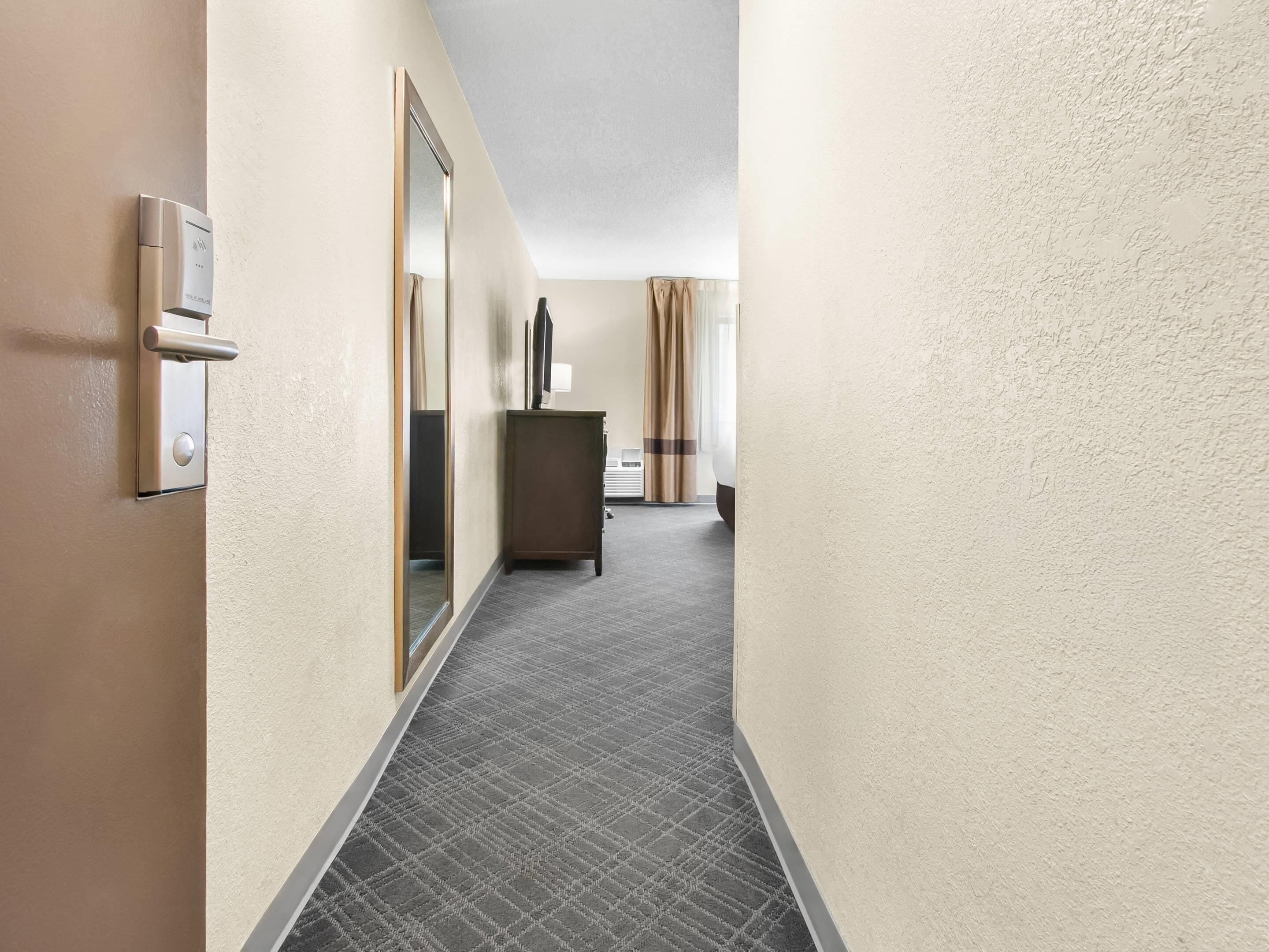 A hotel hallway with a door, a mirror, a cabinet, and a window with curtains.