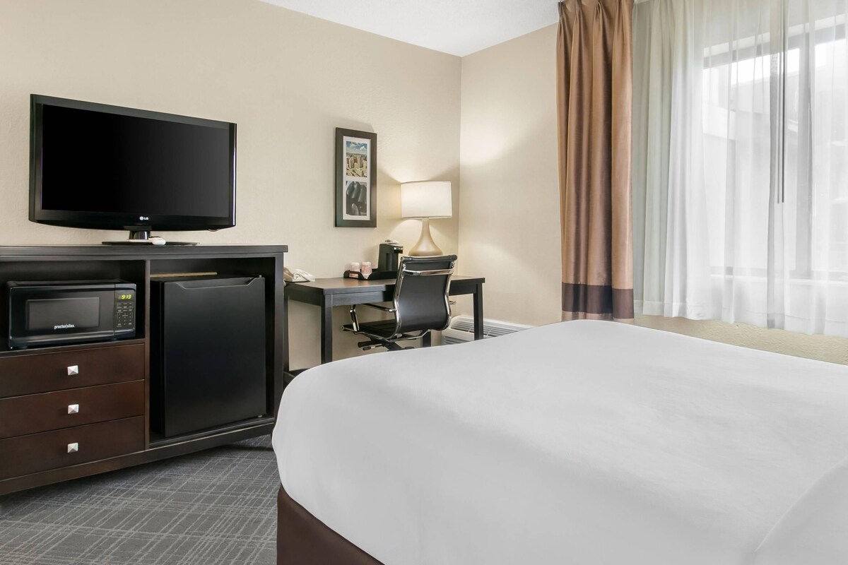Habitación de hotel con cama, escritorio con silla, televisor, microondas, refrigerador, lámpara y ventana con cortinas.