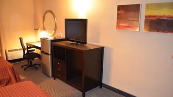 A hotel room with a bed, a desk with a computer, a chair, a refrigerator, and two paintings on the wall.