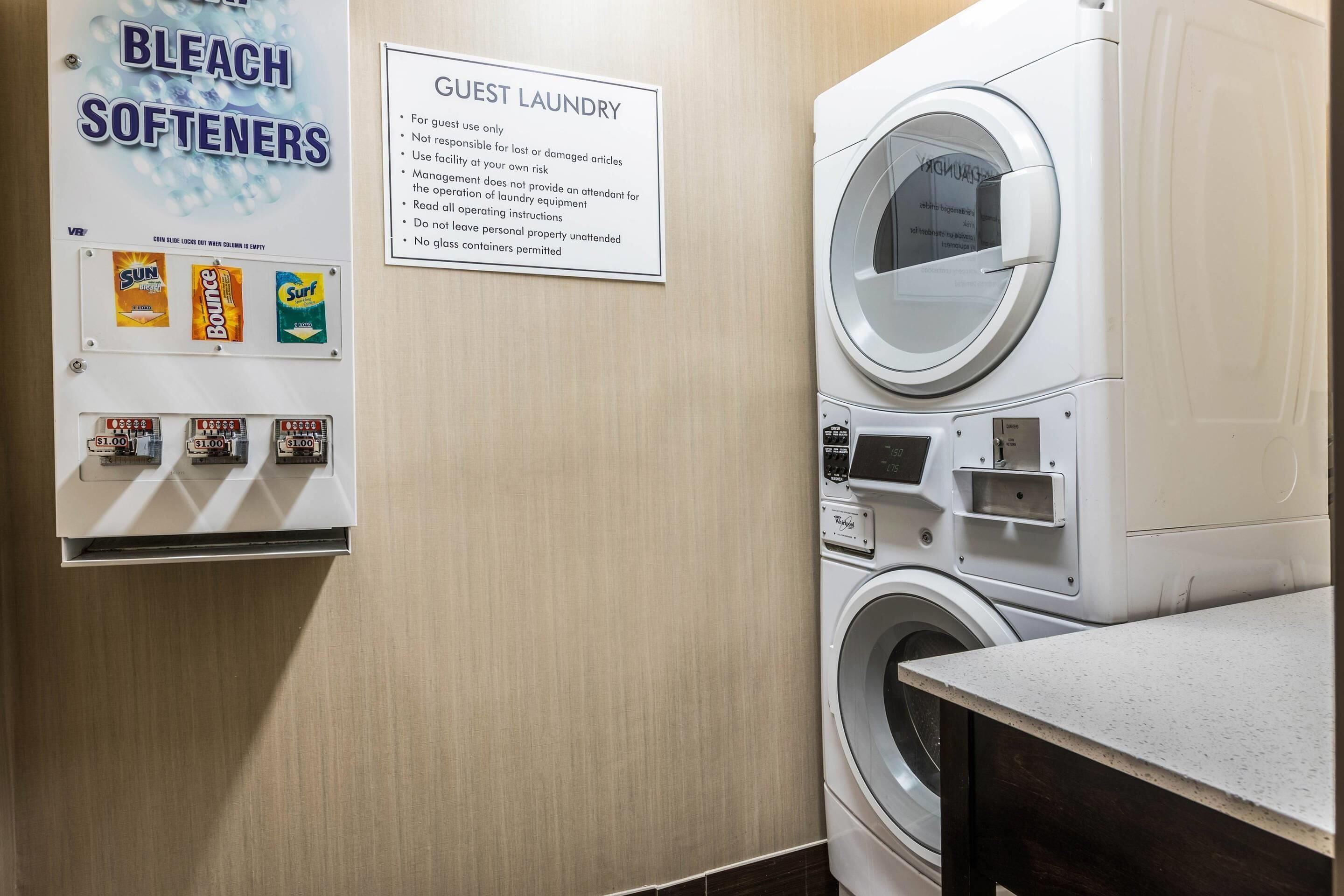 A guest laundry area with a sign detailing usage rules, a wall-mounted detergent dispenser, and a front-loading washing machine.