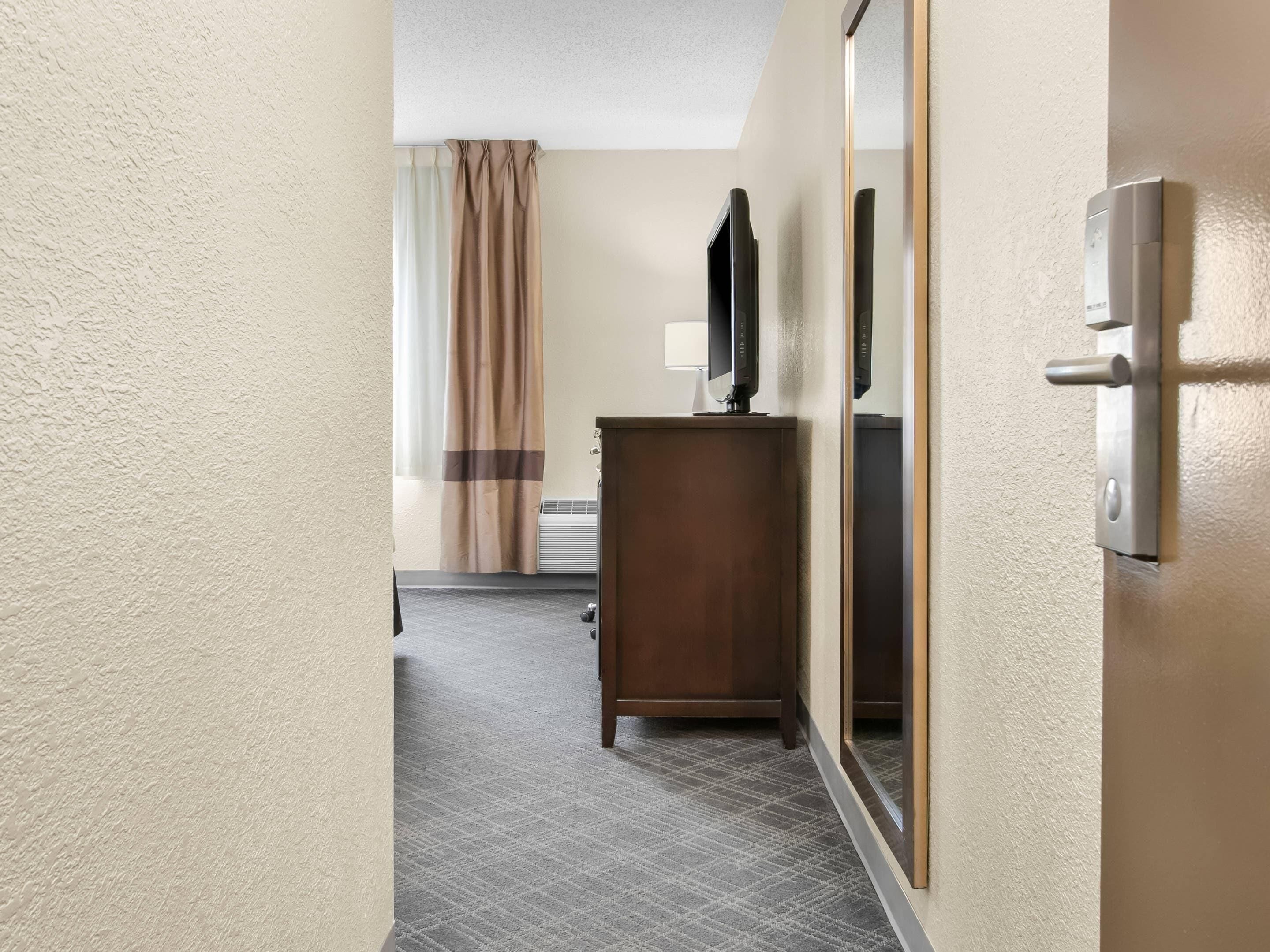 A hotel hallway with a wooden desk, a flat-screen TV, a mirror, and a door with a handle.