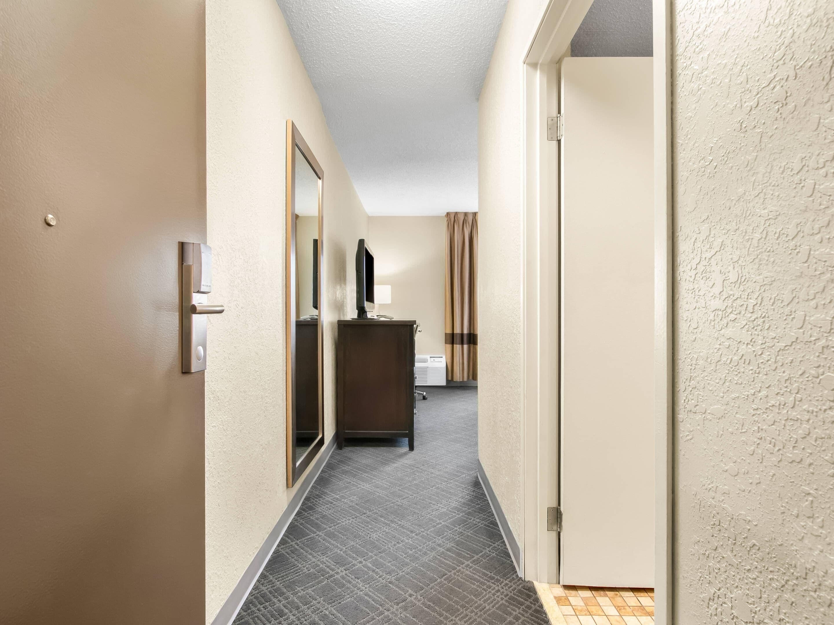 A hotel hallway with a mirror, a desk with a computer, and a window with curtains.