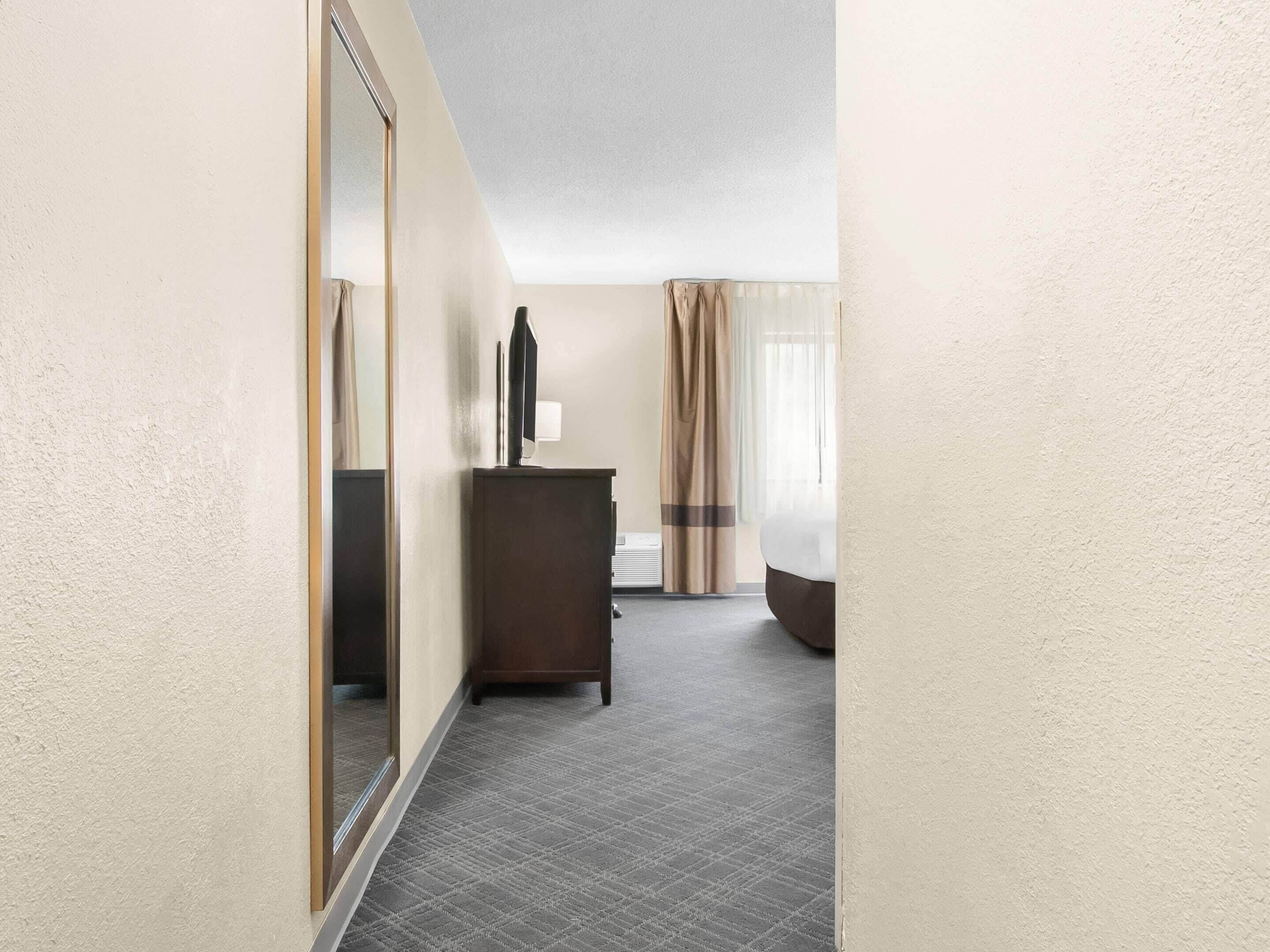 A hotel room with a large mirror, a dresser, a bed, and a window with curtains.