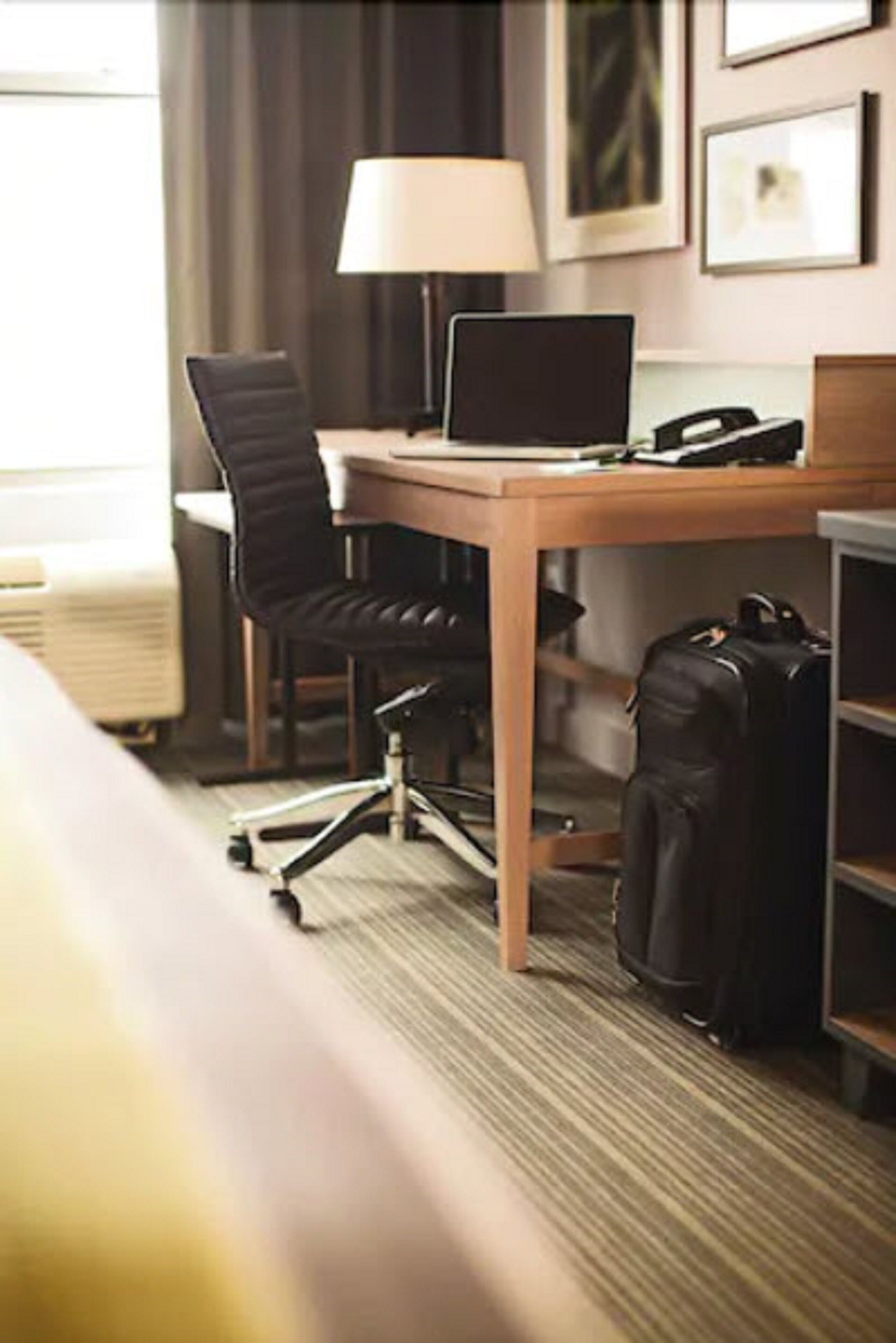 A hotel room with a wooden desk, a laptop, a chair, a lamp, and a suitcase.