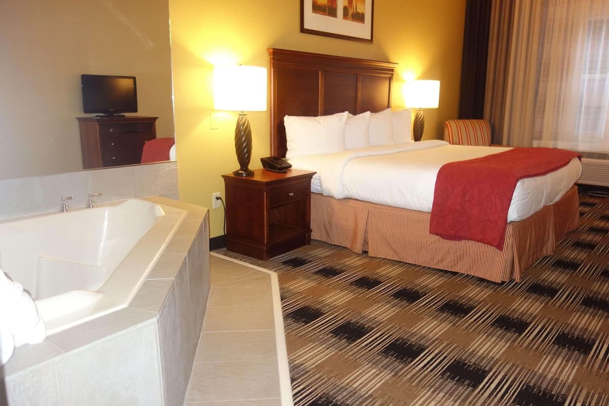 A hotel room with a large bed, two bedside lamps, a television on a dresser, a bathtub, and a window with curtains.
