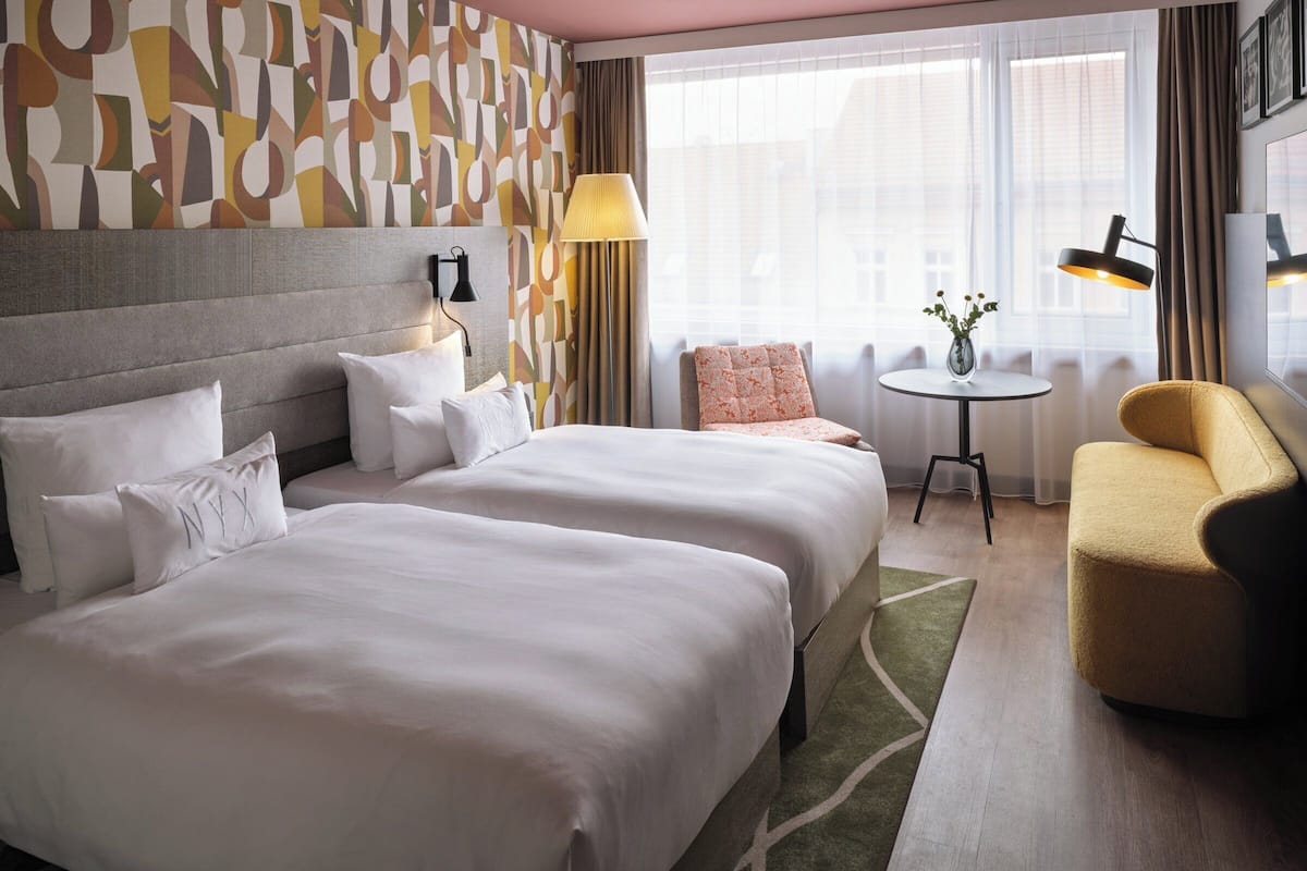 Una habitación de hotel con dos camas, una mesita con una lámpara, una silla y un ventanal con cortinas.