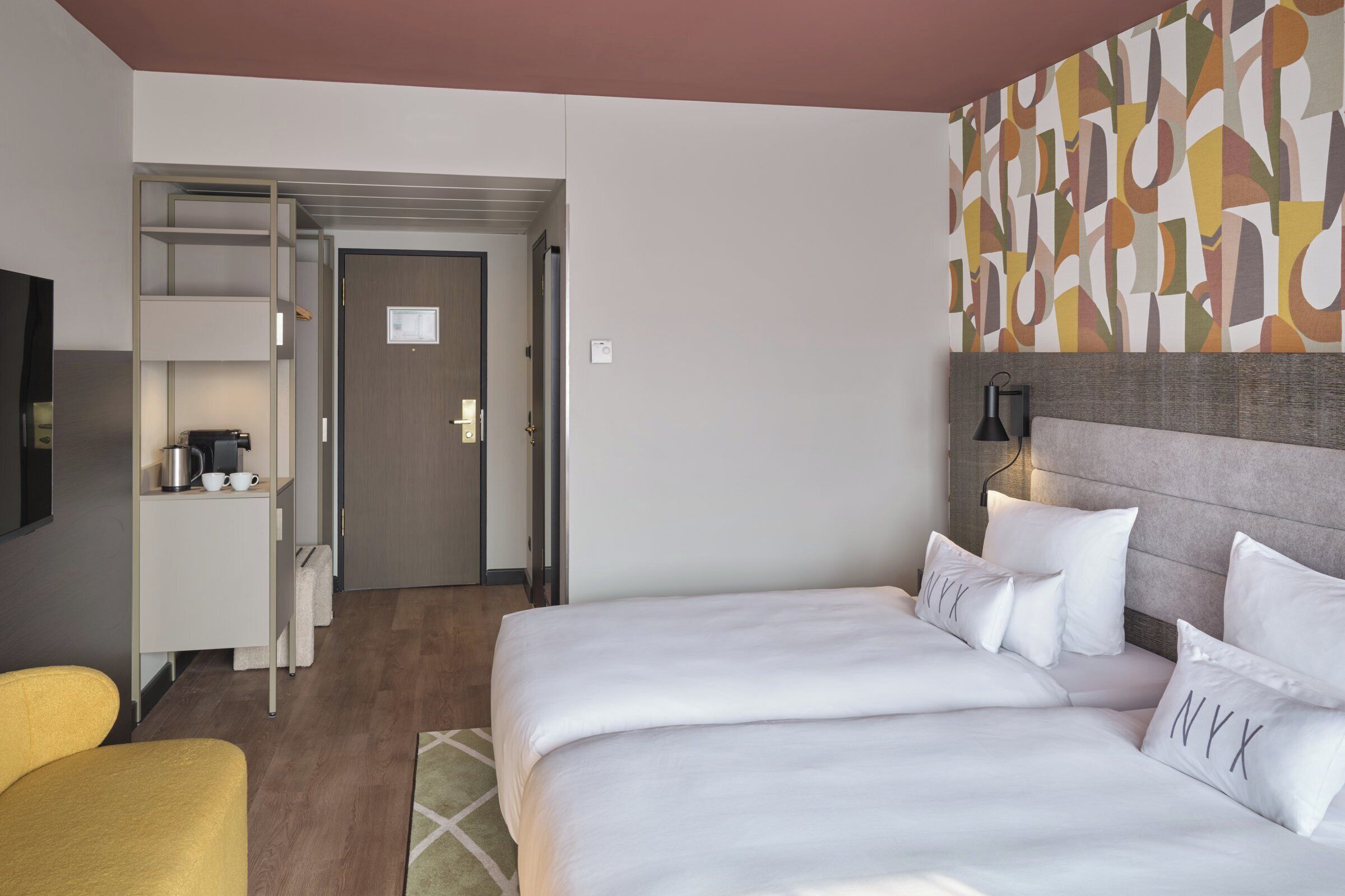Quarto de hotel com cama, escrivaninha, cadeira, televisão e parede com arte abstrata.