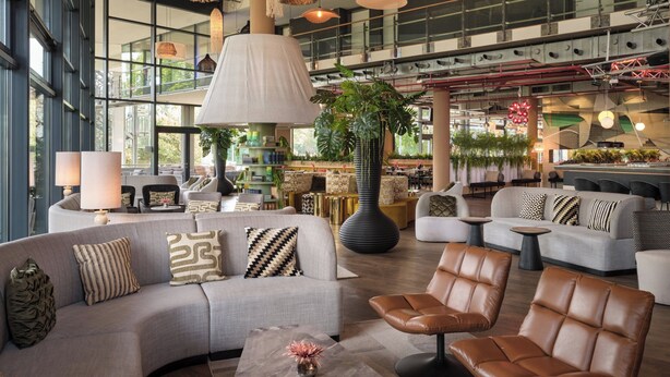 Un vaste espace de détente avec des canapés, des fauteuils et une table basse, agrémenté d’une grande plante en pot et de lampes suspendues.