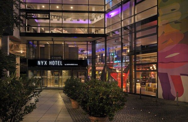 The NYX HOTEL by LEONARDO Hotels with a modern glass facade, a visible entrance, and a colorful mural on the right side.
