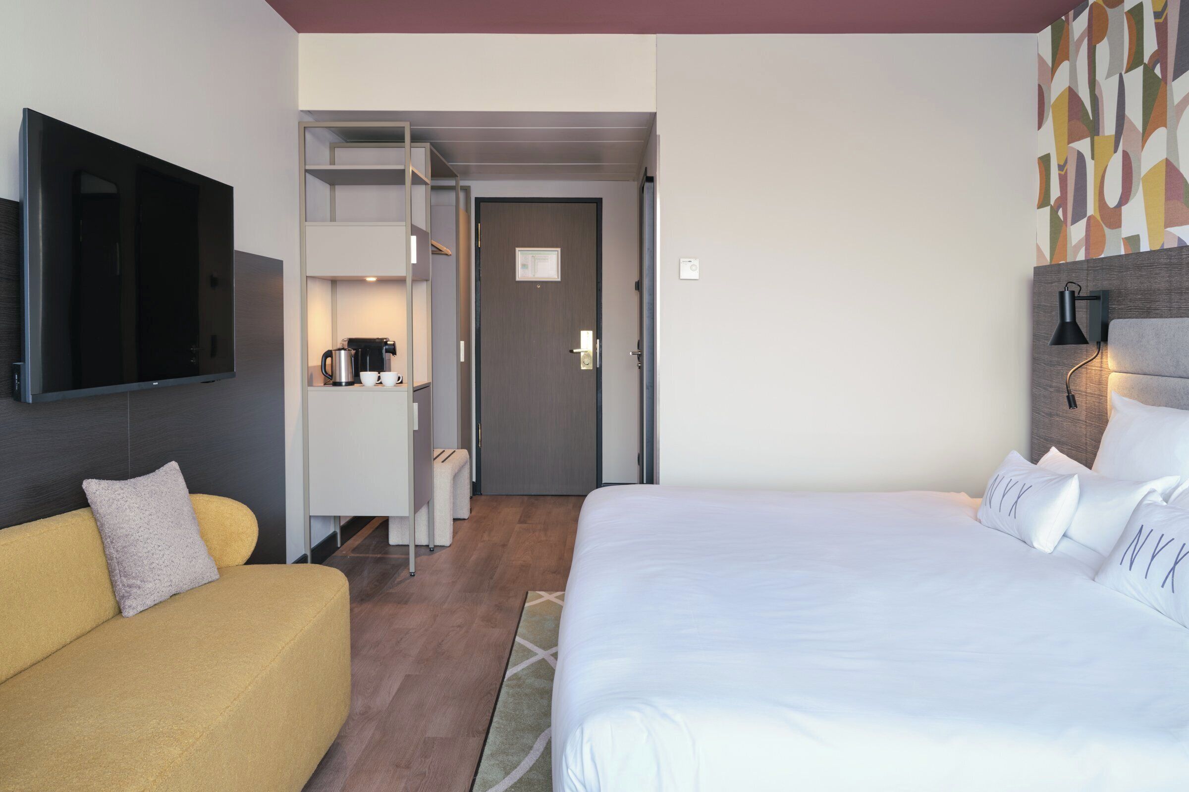 A hotel room with a bed, a sofa, a TV, a coffee machine, and a wardrobe.