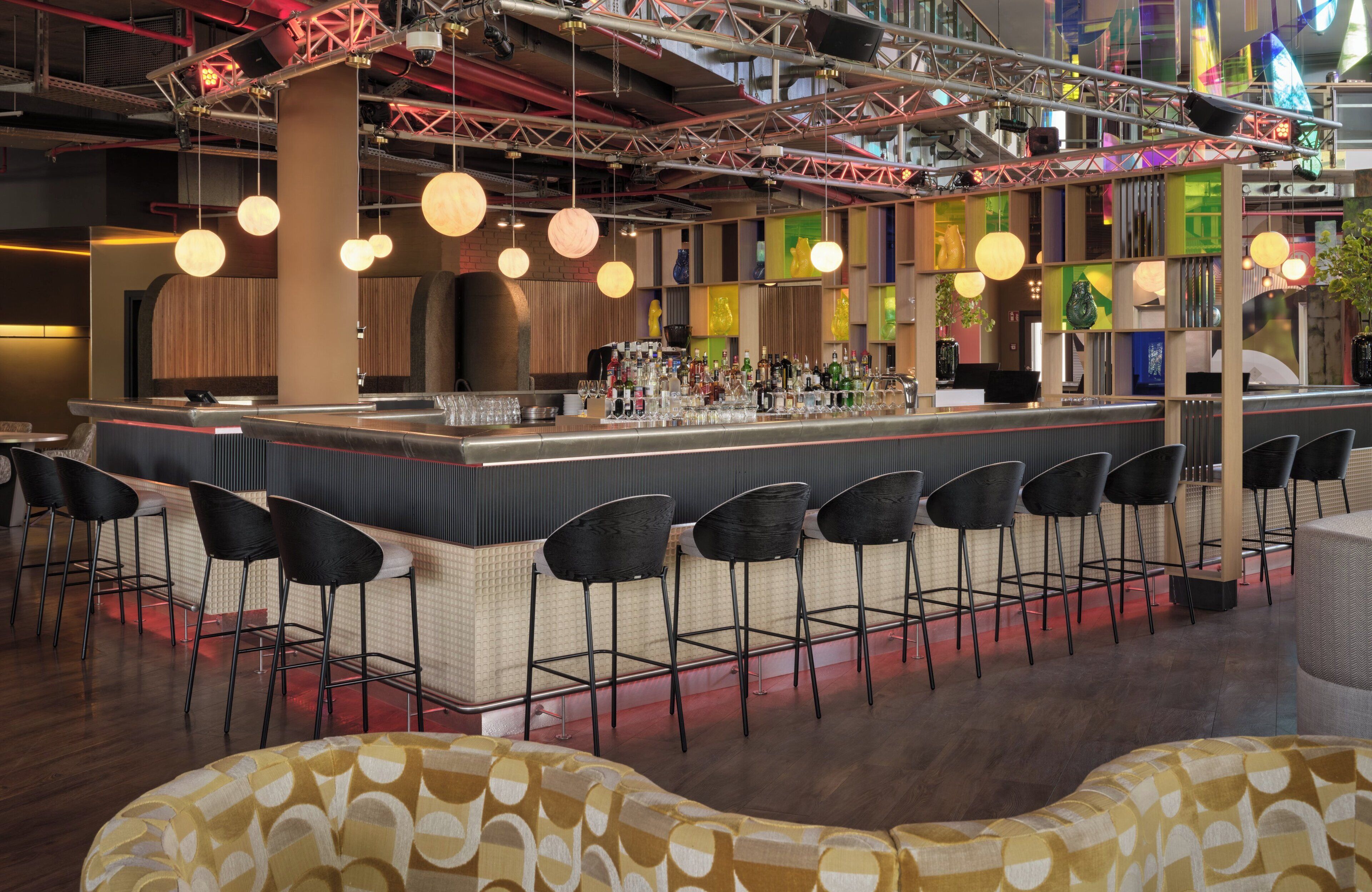 A modern bar area with a central counter, multiple bar stools, hanging spherical lights, and a variety of colorful wall decorations.