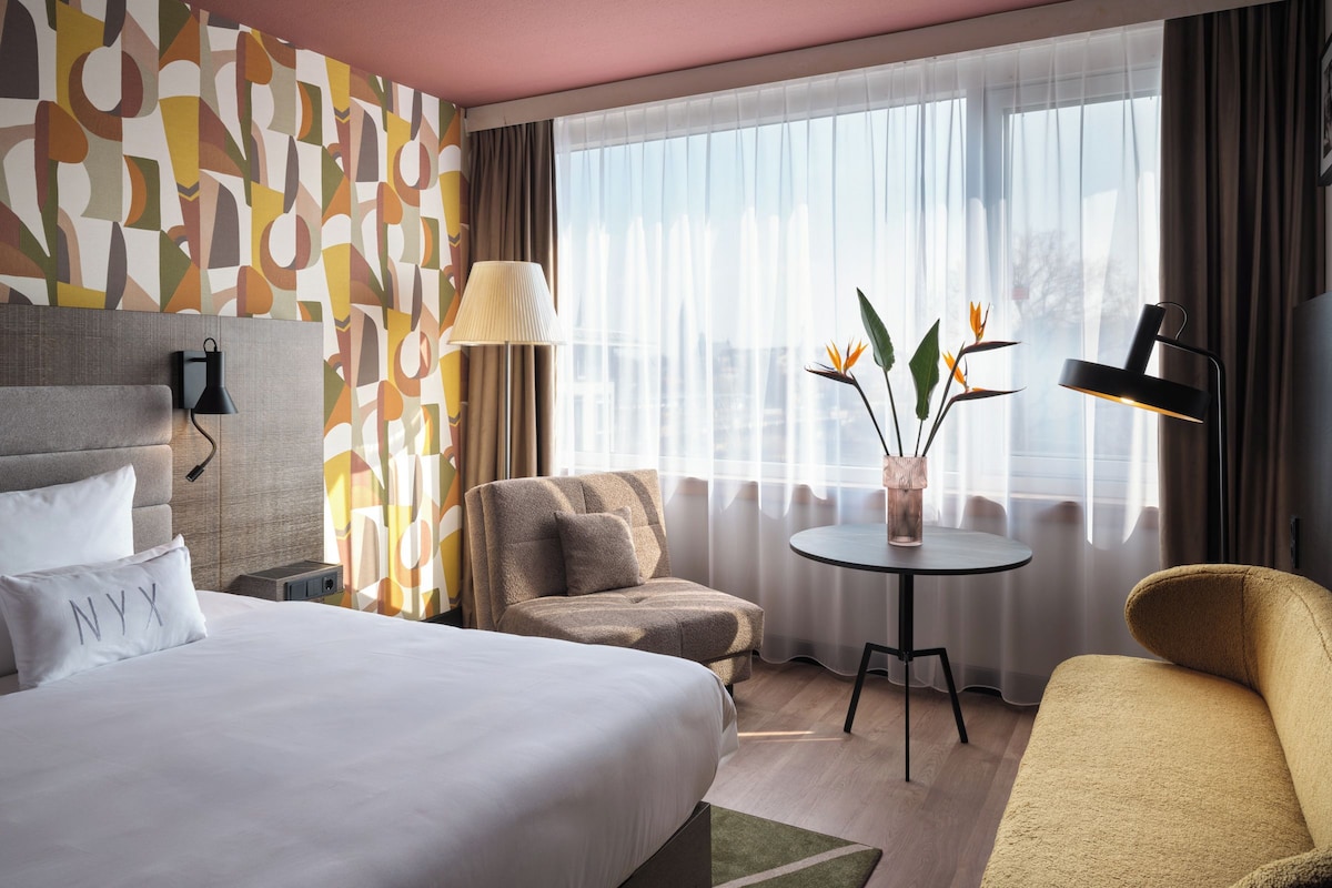 Quarto de hotel com cama, sofá, poltrona, mesa pequena e janela com cortinas.