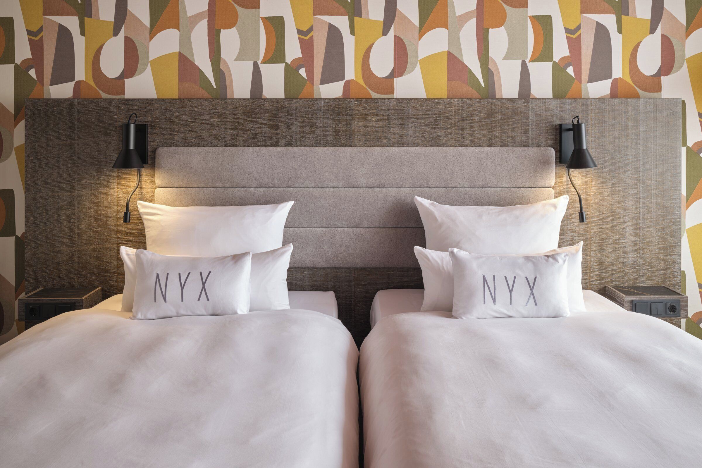 Un lit double avec deux oreillers portant l’inscription « NYX » et une tête de lit agrémentée de deux lampes fixées au mur.