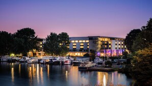 Un port de plaisance avec des bateaux amarrés, un hôtel aux fenêtres illuminées et une étendue d’eau calme qui reflète les lumières.