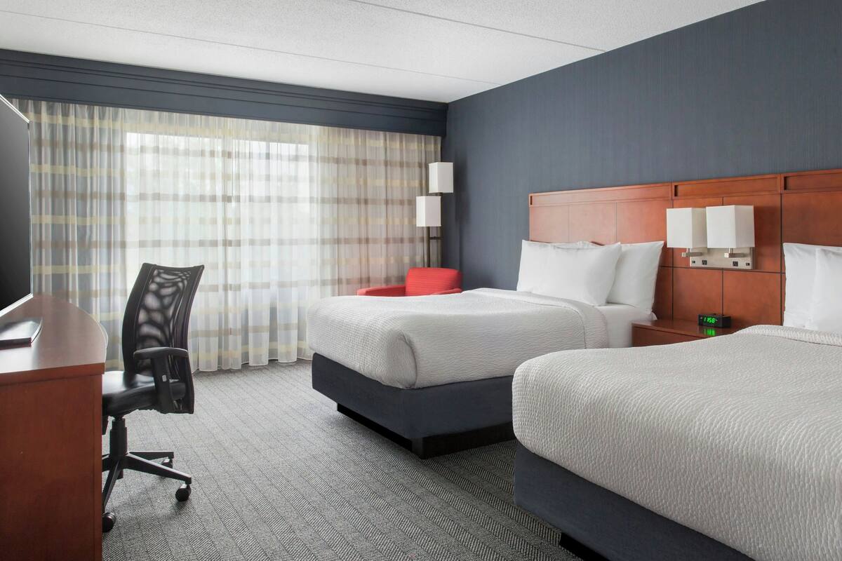 A hotel room with two beds, a desk, a chair, and a nightstand with lamps.