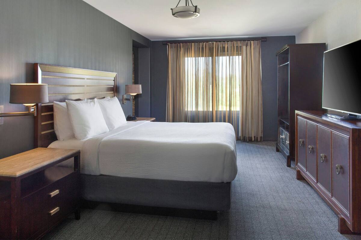 A hotel room with a large bed, a wooden nightstand, a dresser, a television, and a window with curtains.