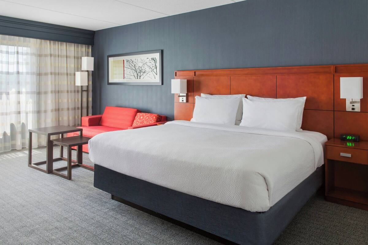 A hotel room with a large bed, a small table, a red chair, and a nightstand with a clock.