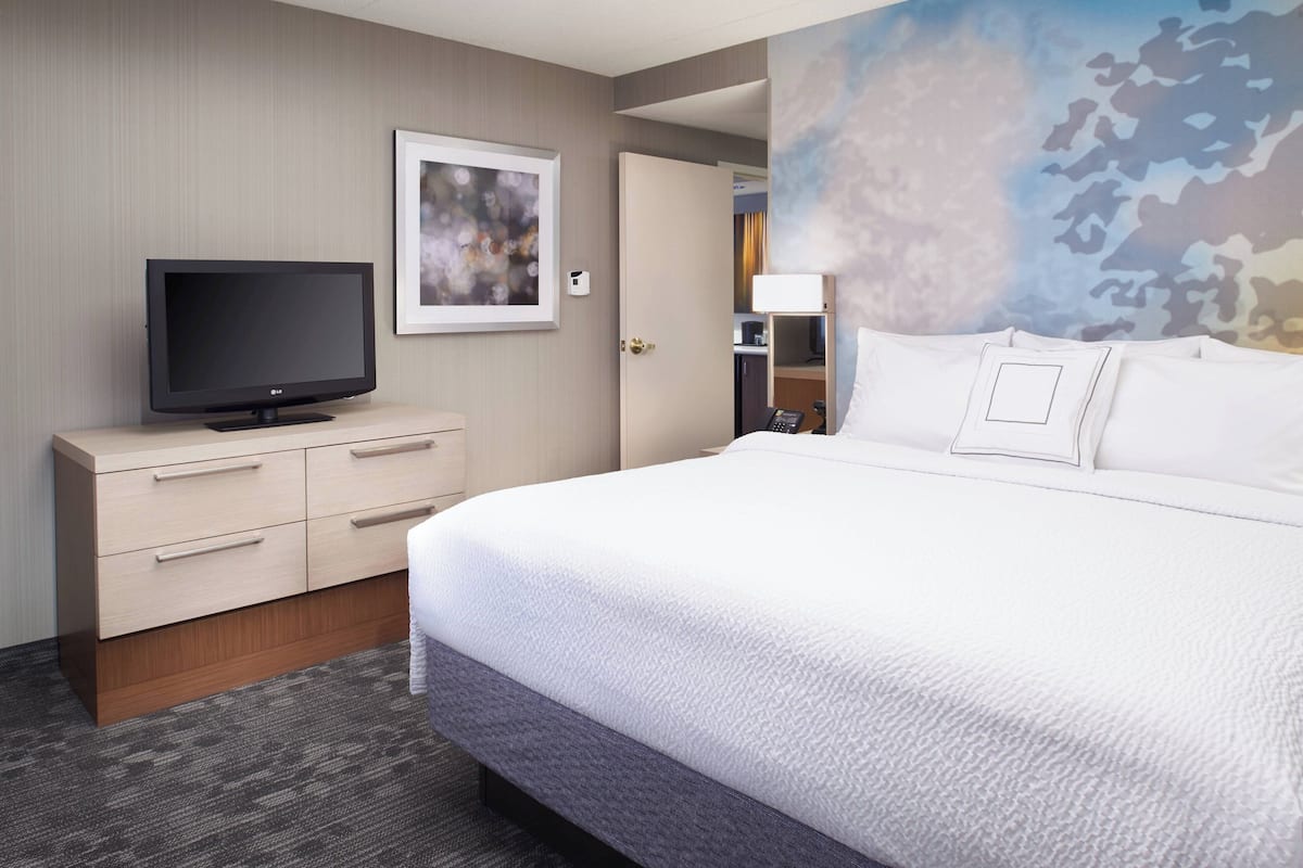 Quarto de hotel com uma cama, uma televisão em uma cômoda de madeira, um quadro na parede e uma porta que dá para outro cômodo.
