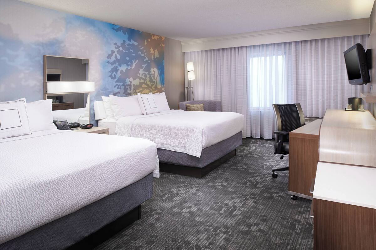 Quarto de hotel com duas camas, uma escrivaninha, uma cadeira, uma televisão e uma janela grande com cortinas.