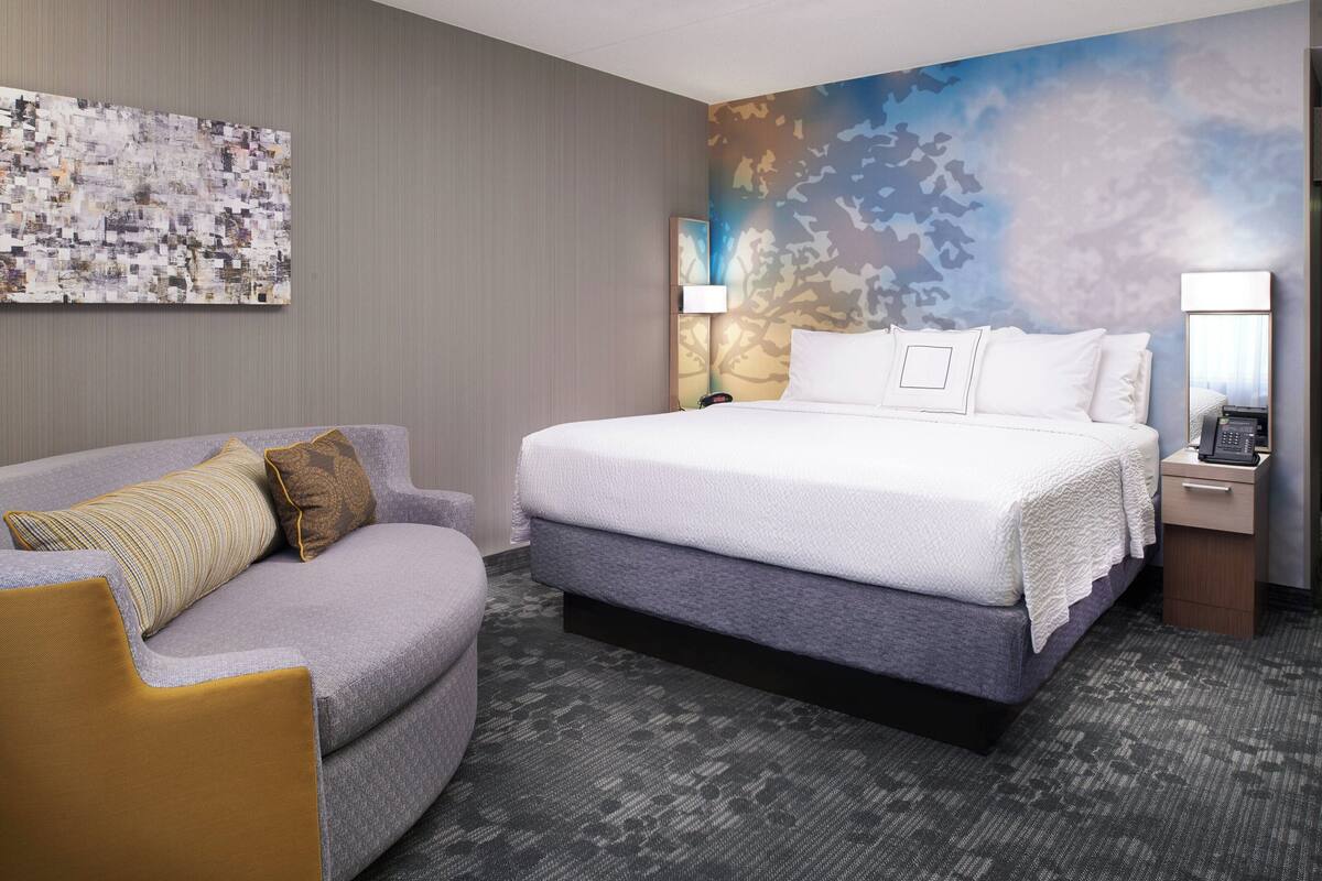Quarto de hotel com uma cama grande, um sofá, uma mesa de cabeceira com luminária e um mural de paisagem urbana na parede.