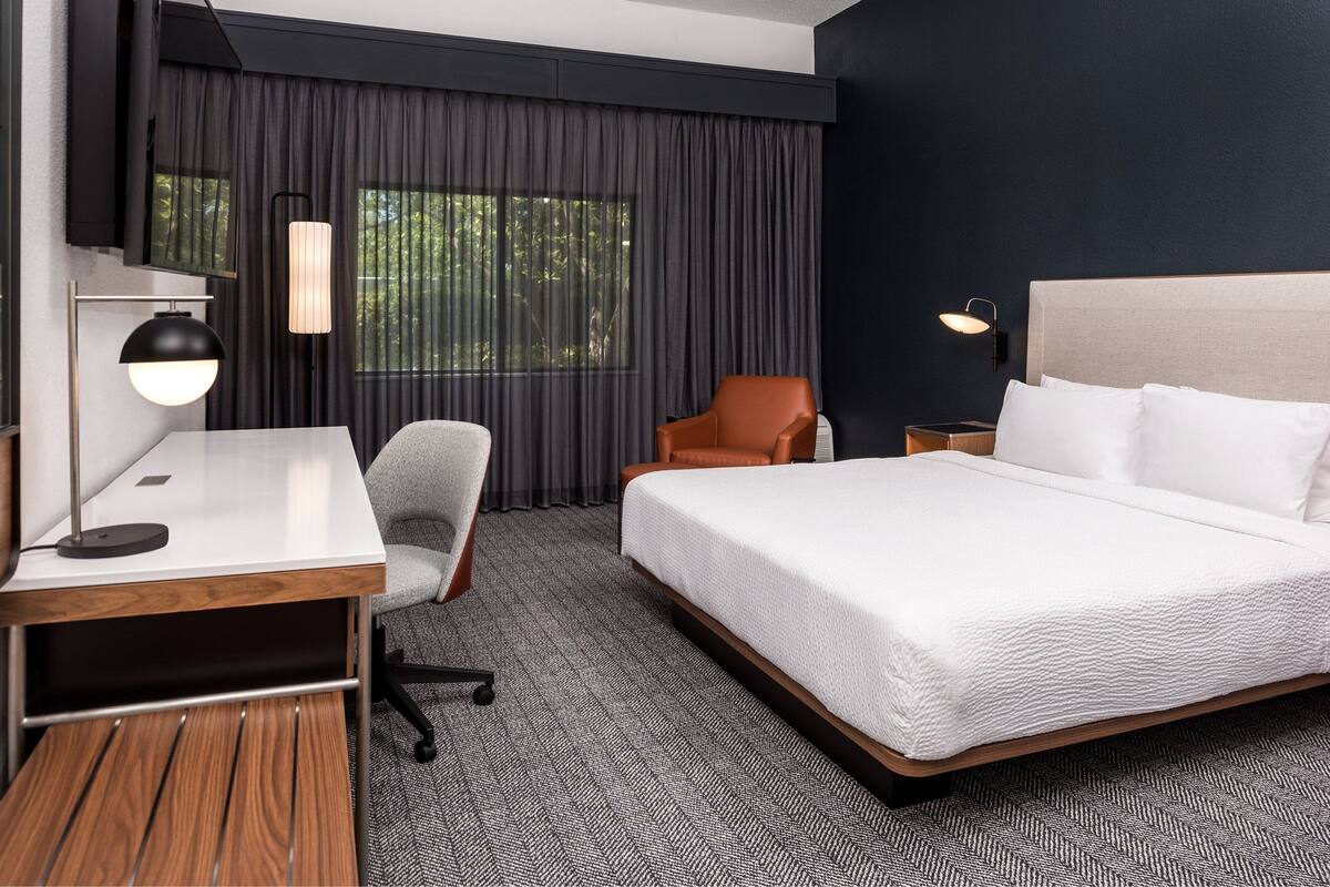 A hotel room with a large bed, a desk with a lamp, a chair, and a view of trees through the window.