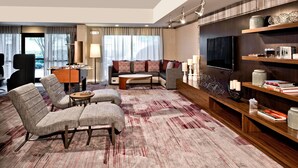A modern hotel room with a sofa, armchairs, a coffee table, a TV, and a bookshelf. - Courtyard by Marriott Ballantyne (Charlotte)