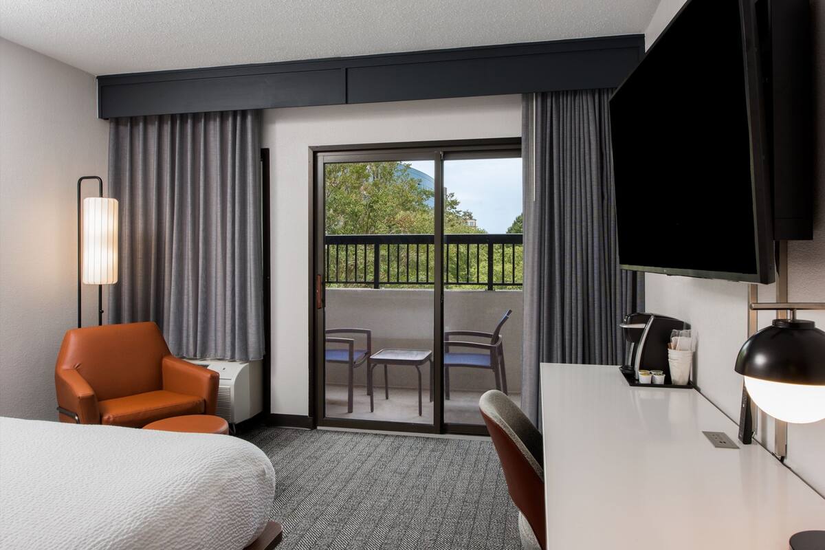 A hotel room with a bed, a desk, a chair, a TV, a lamp, and a balcony with a view of greenery.