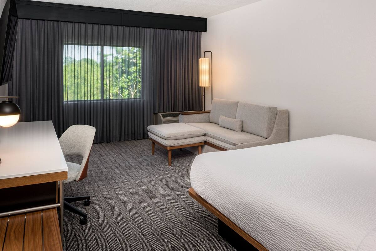 A hotel room with a bed, a desk, a chair, a sofa, and a large window with curtains.