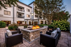 Un patio exterior con una chimenea de piedra, mobiliario de mimbre y un edificio con balcones y ventanas amplias.