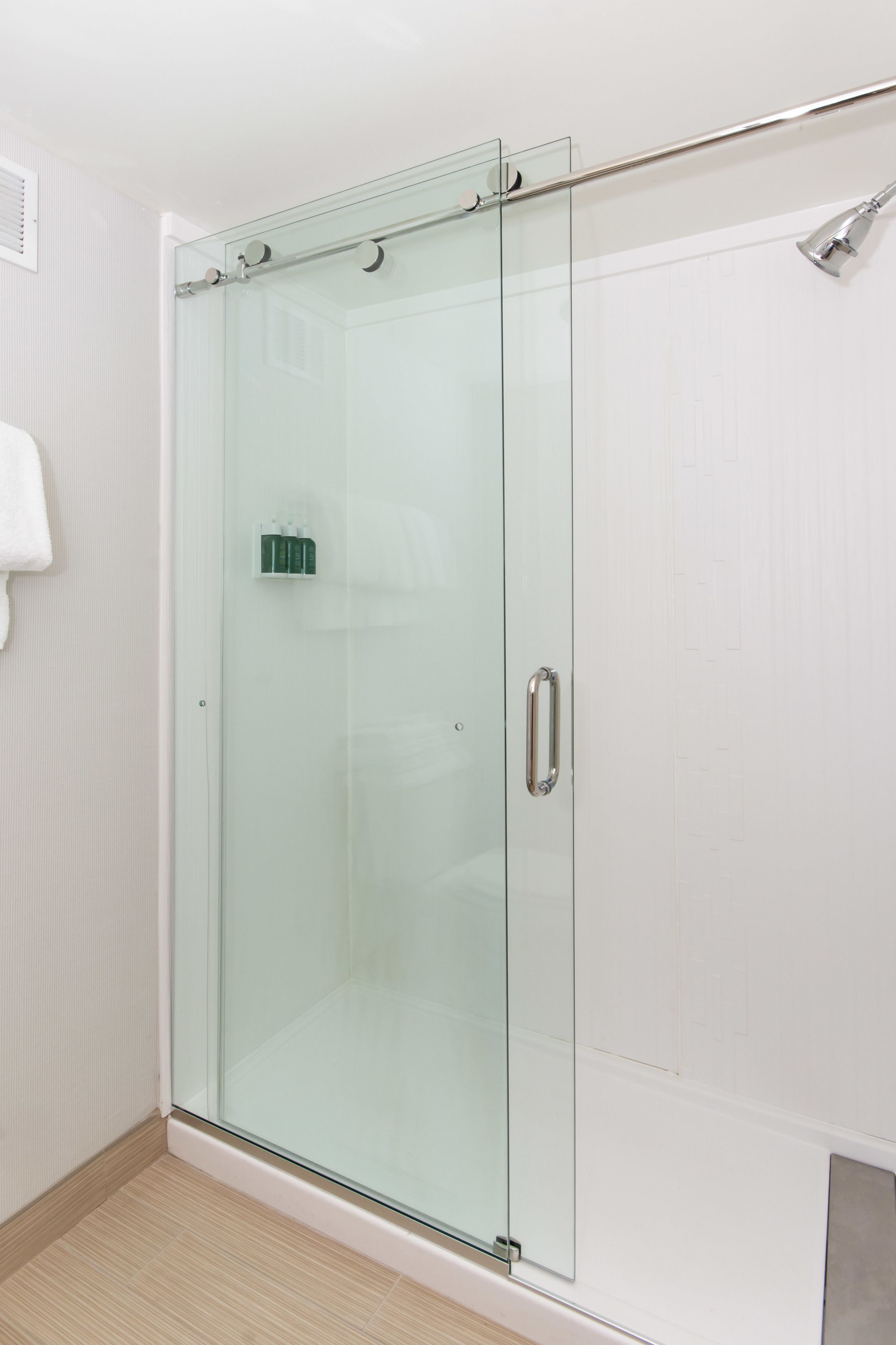 Een douche met glazen wand, een witte betegelde muur en een lichtgekleurde vloer.