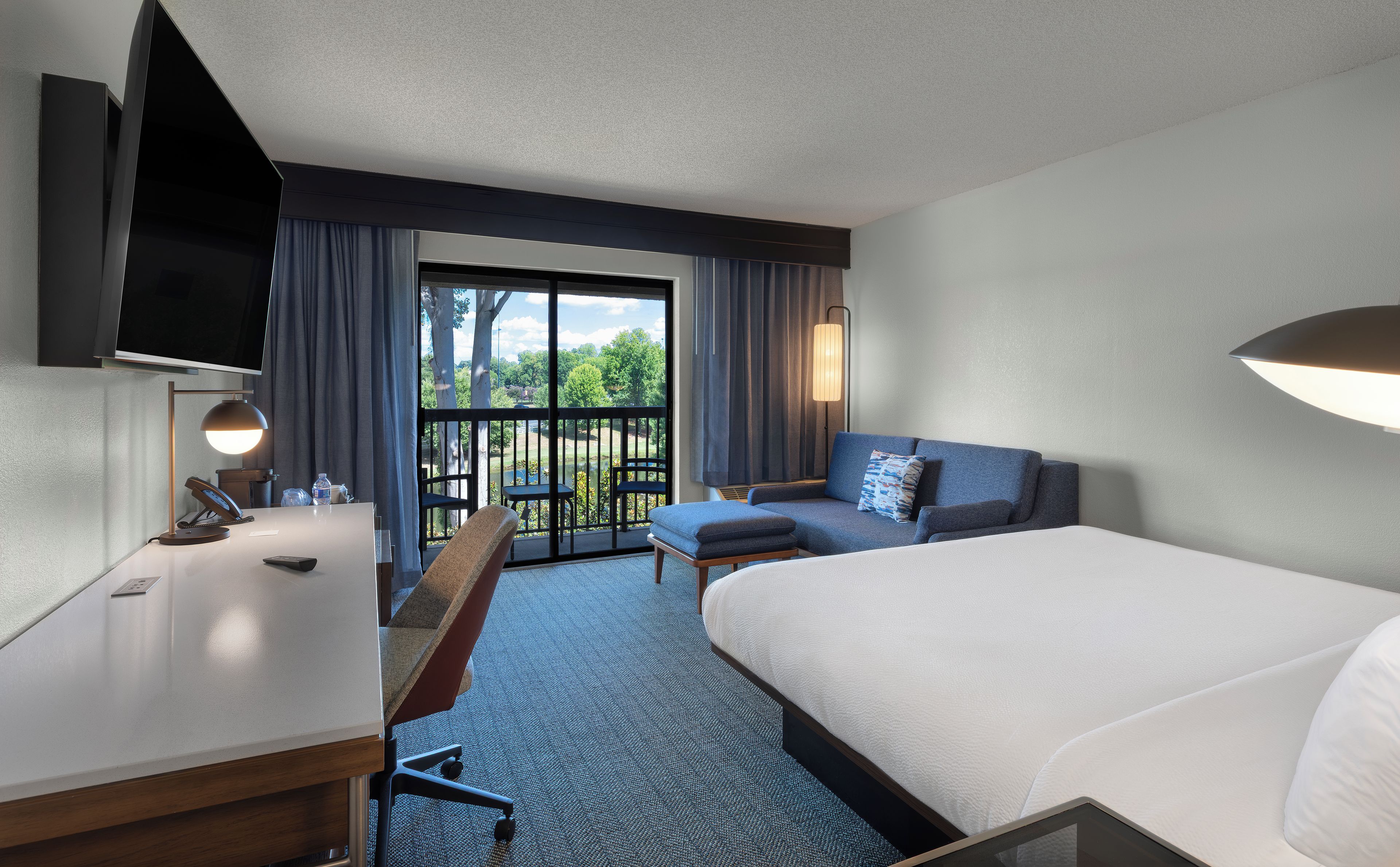 A hotel room with a bed, a desk with a telephone, a chair, a sofa, and a large window offering a view of trees and a building.