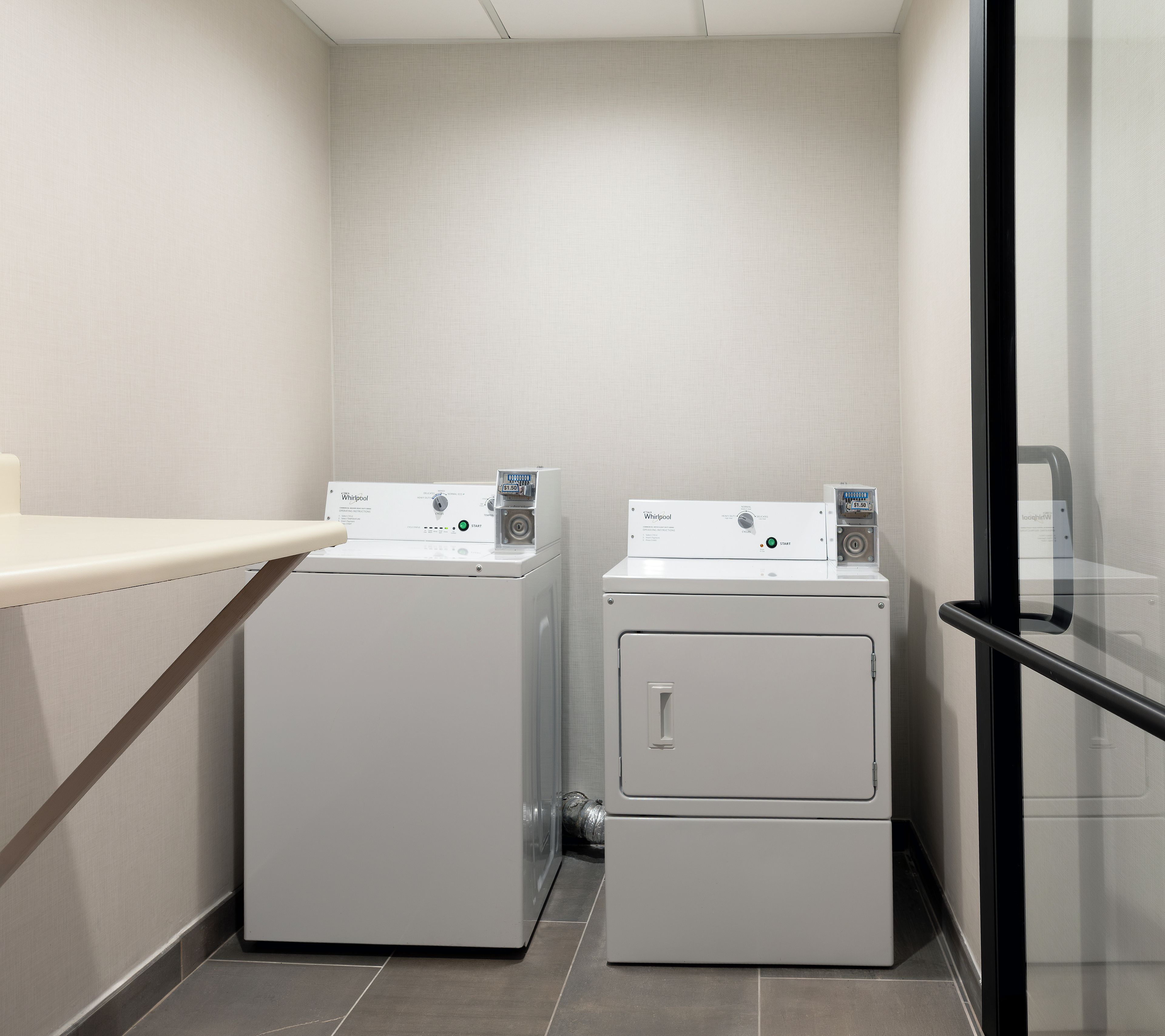 A laundry room with two white Whirlpool washers and dryers, a small countertop, and a glass door.