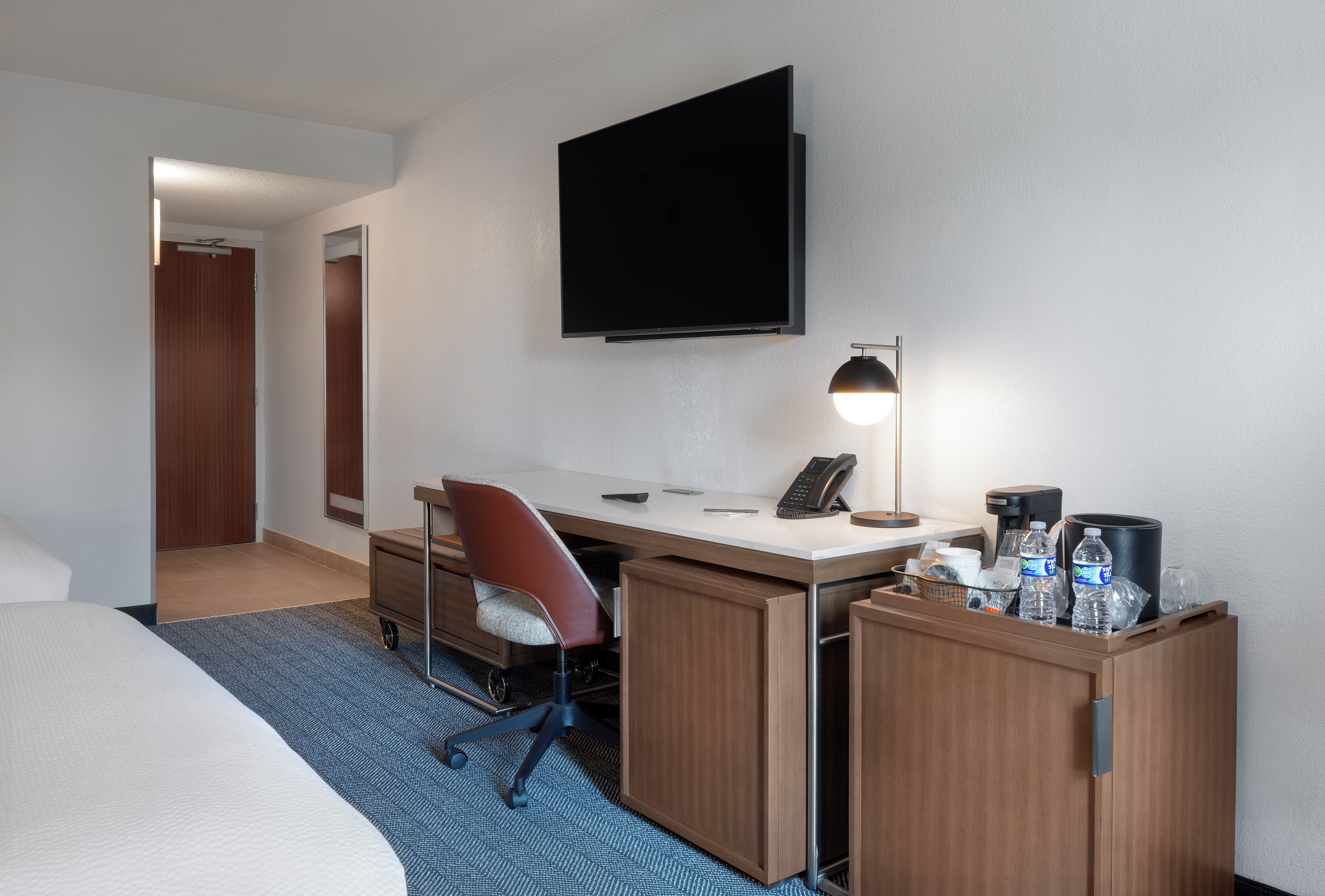 A hotel room with a bed, a desk with a chair, a flat-screen TV, a desk lamp, a telephone, and a minibar.