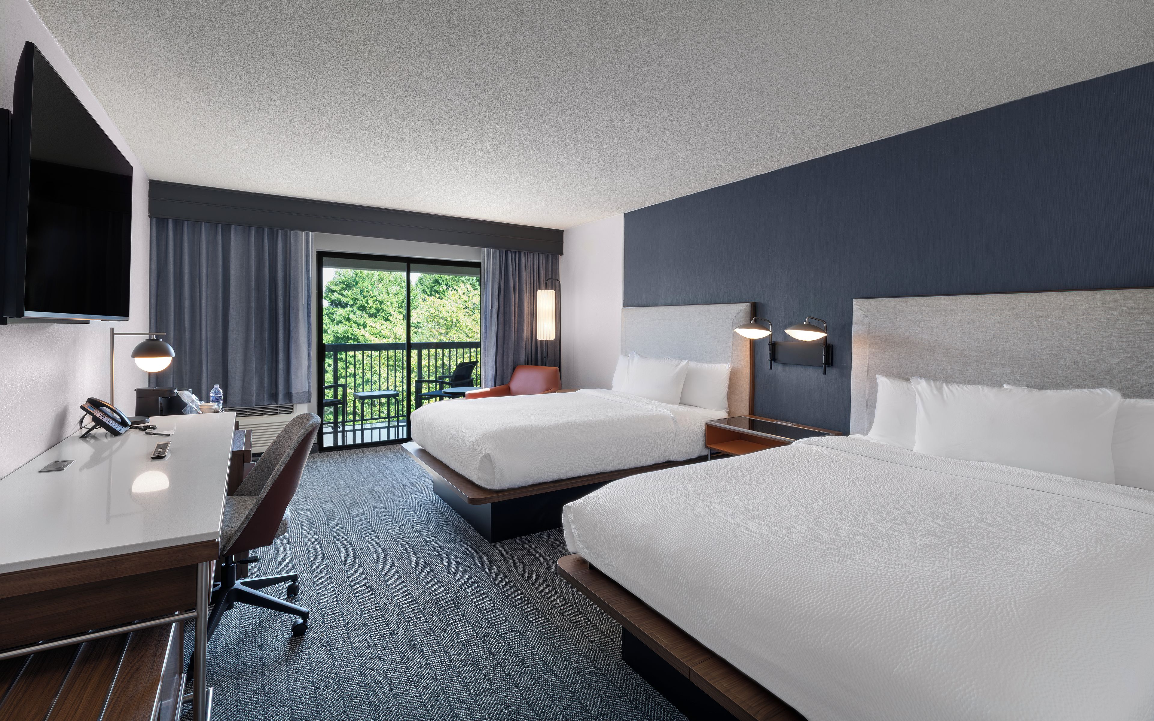 A hotel room with two beds, a desk, a chair, a telephone, and a view of greenery through the window.