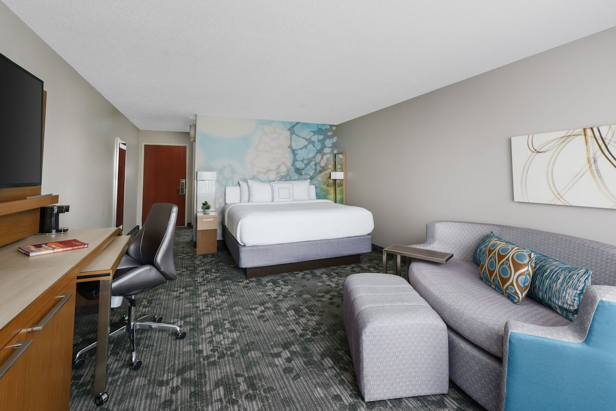 A hotel room with a bed, a desk with a chair, a sofa, a small table, and a wall with a painting.