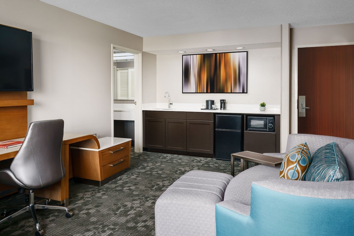 A modern hotel room with a grey sofa, a wooden desk, a flat-screen TV, a kitchenette, and a large abstract painting on the wall.