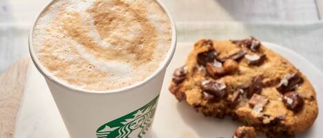 A Starbucks cup with a frothy top and a cookie with chocolate chips on a white plate.