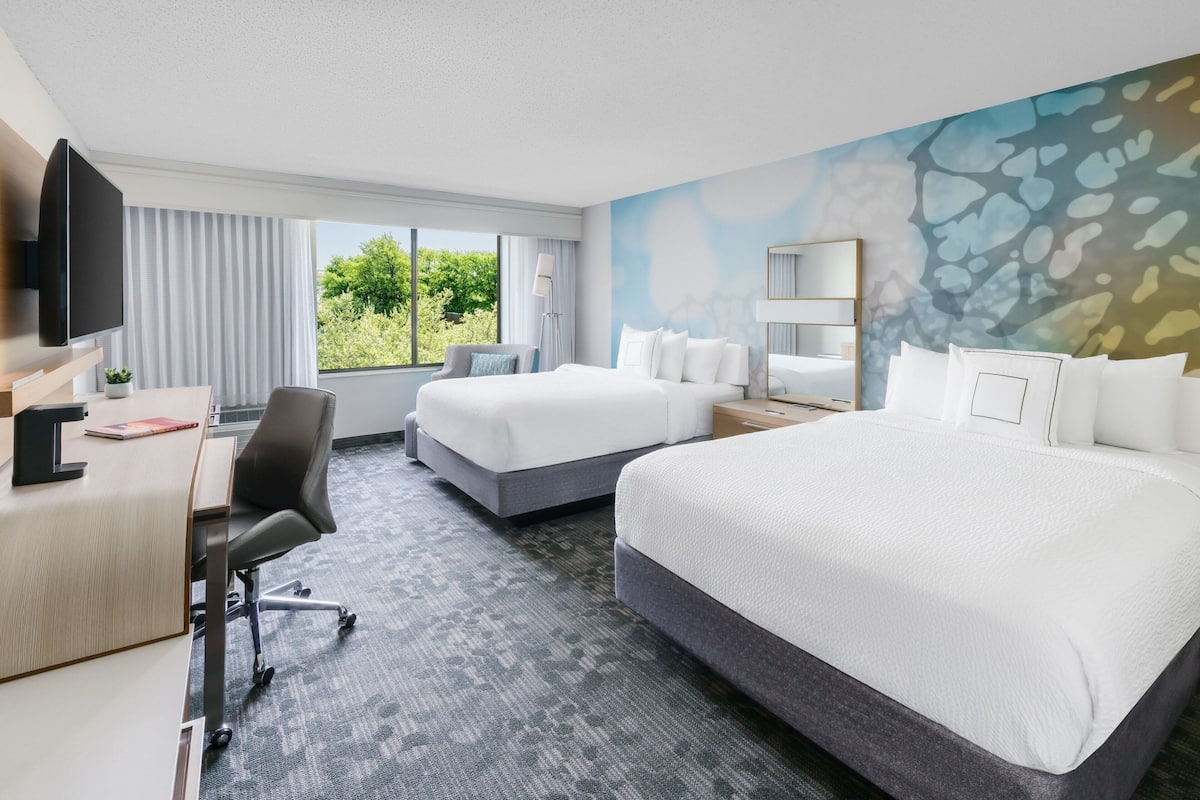 A hotel room with two beds, a desk with a chair, a TV, and a large window with curtains.