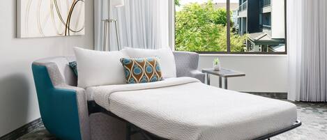 A modern hotel room with a single bed, a view of greenery outside, a small table, and a floor lamp.