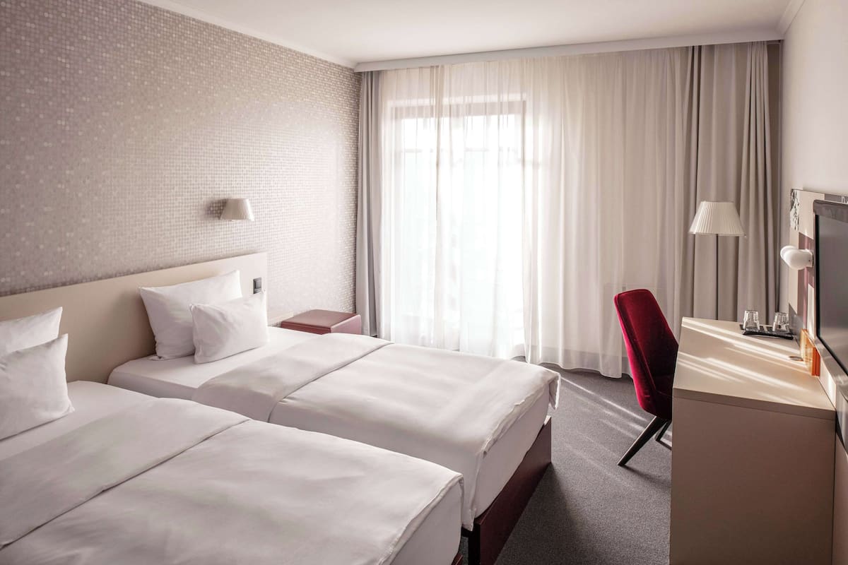 A hotel room with two beds, a desk, a chair, a TV, and a window with curtains.