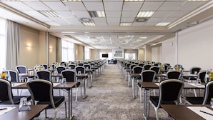 Un'ampia sala conferenze con file di tavoli e sedie, un palco nella parte anteriore e uno schermo di grandi dimensioni.