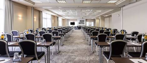 A large conference room with rows of tables and chairs, a stage at the front, and a large screen.