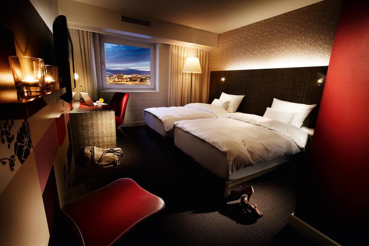 A hotel room with a bed, a desk, a chair, a window with a view, and a red wall.