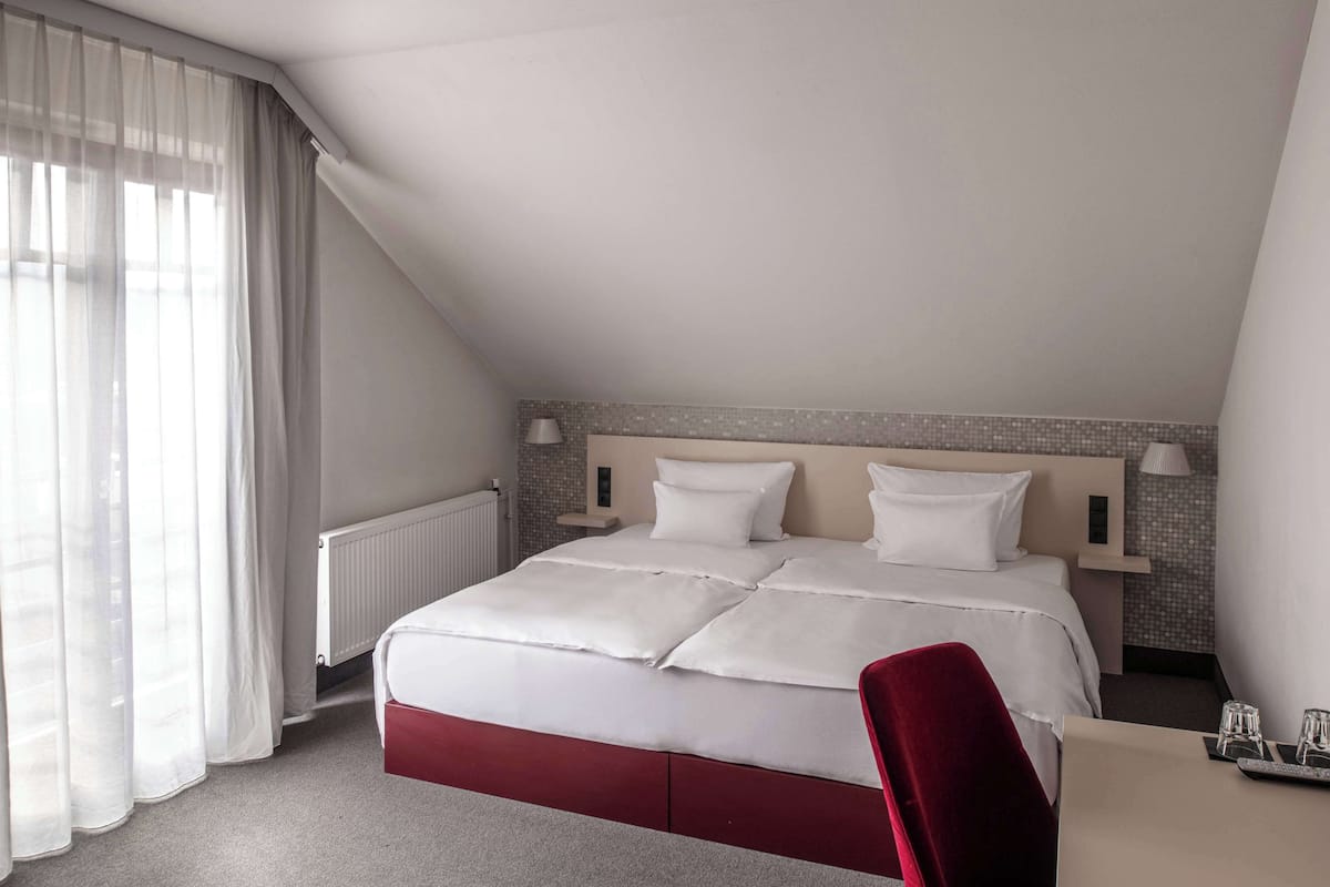 A hotel room with a large bed, a red chair, a small table with glasses, and a window with sheer curtains.