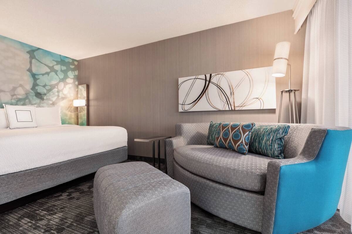 A hotel room with a bed, a grey sofa with a blue accent, a small table, and a wall with abstract art.