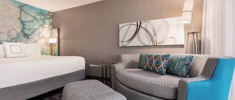 A hotel room with a bed, a grey sofa with a blue accent, a small table, and a wall with abstract art.